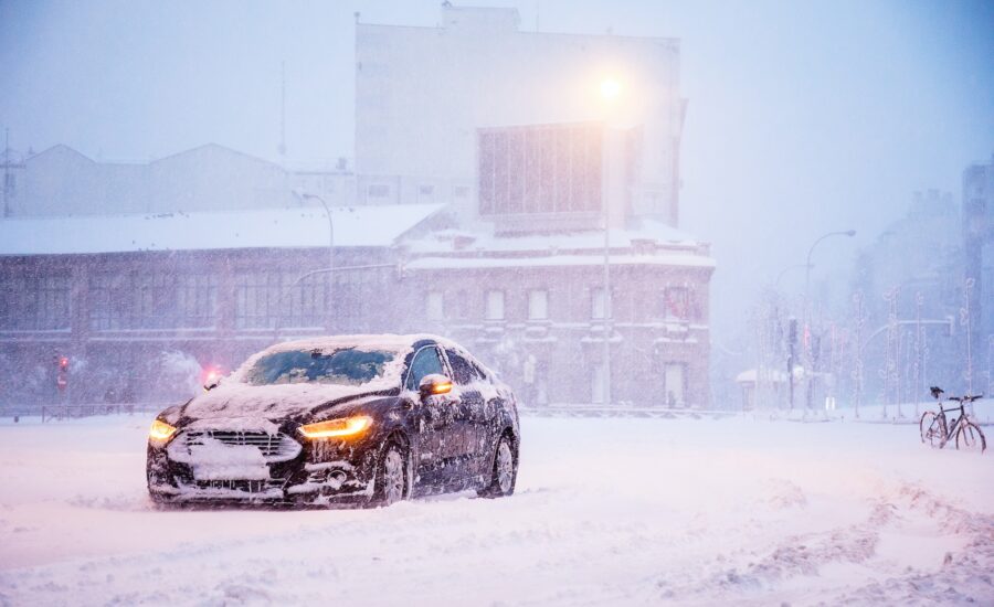 Ein schwarzes Auto steht vor einem großen Industriegebäude. Es schneit und das Auto hat die Scheinwerfer angeschaltet.